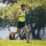 Full length of young bearded sportsman in activewear running on grassy lawn in city park near green trees during outdoor workout